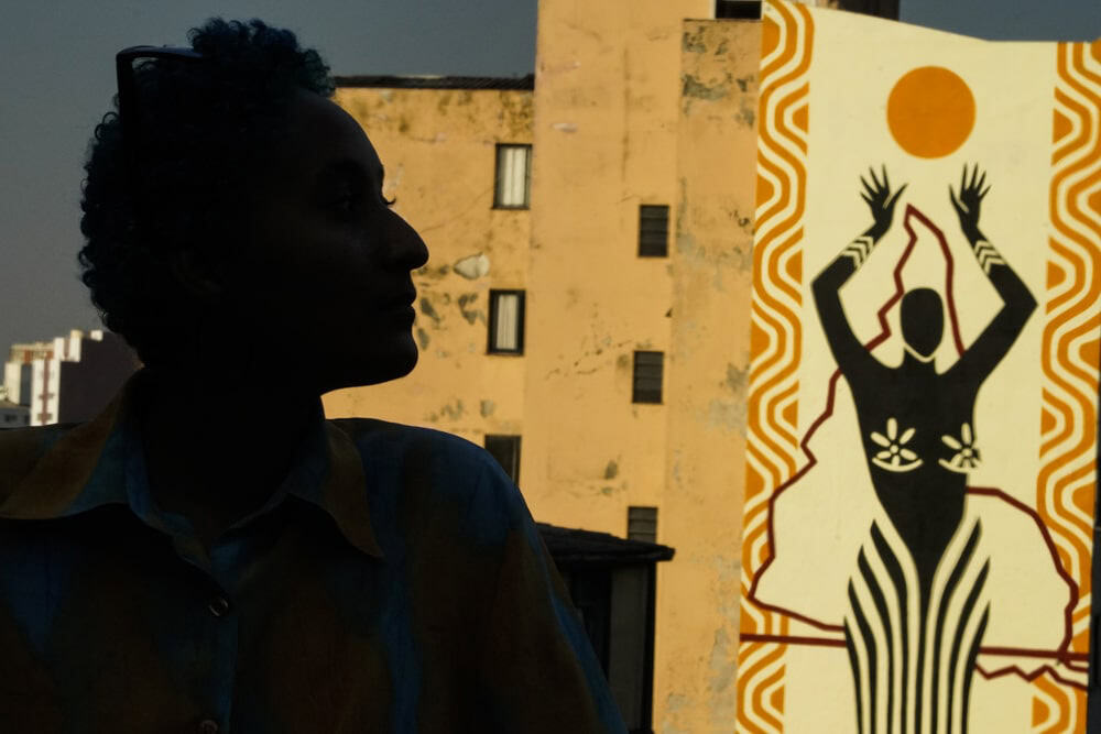 A person with short curly hair sits against an urban backdrop